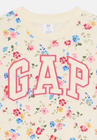 Krēmveida t-krekls ar ziedu rakstu, kas rotāts ar lielu, rozā "GAP" logotipu, ar apaļu dekoltē un mīkstu, teksturētu audumu.