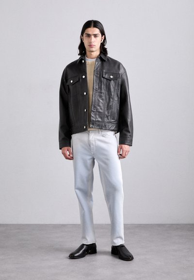 Black leather jacket with two front pockets, gray sweater, light blue jeans, and black shoes. Model stands centrally against a light background.