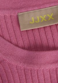 Tejido de suéter de punto rosa con textura acanalada. Cuenta con una etiqueta beige que muestra "JJXX" en letras amarillas.