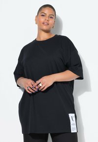 Black oversized t-shirt made of cotton, featuring short sleeves and a side tag with white text. Smooth texture, relaxed fit, and simple design.