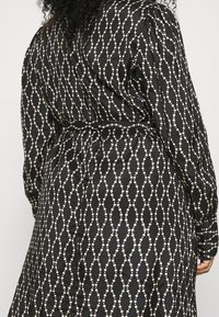 Czarna sukienka z białym wzorem przeplatanych pętli, wykonana z gładkiego materiału, z długimi rękawami i zapinanym w pasie krojem.