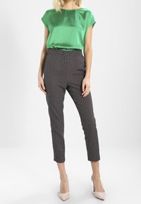 Blusa de satén verde con mangas cortas, combinada con pantalones ajustados negros con estampado. La modelo lleva unos zapatos de tacón nude.