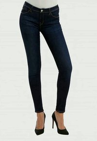 Γυναίκα που φορά σκούρα μπλε στενά τζιν και μαύρες ψηλοτάκουνες γόβες, στέκεται με το ένα πόδι ελαφρώς λυγισμένο.