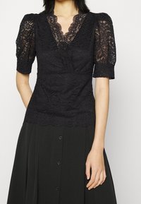 Schwarzes Spitzentop mit tiefem V-Ausschnitt, kurzen Puffärmeln und gewellten Kanten. Kombiniert mit einem schwarzen, knopfverschlossenen, fließenden Rock.