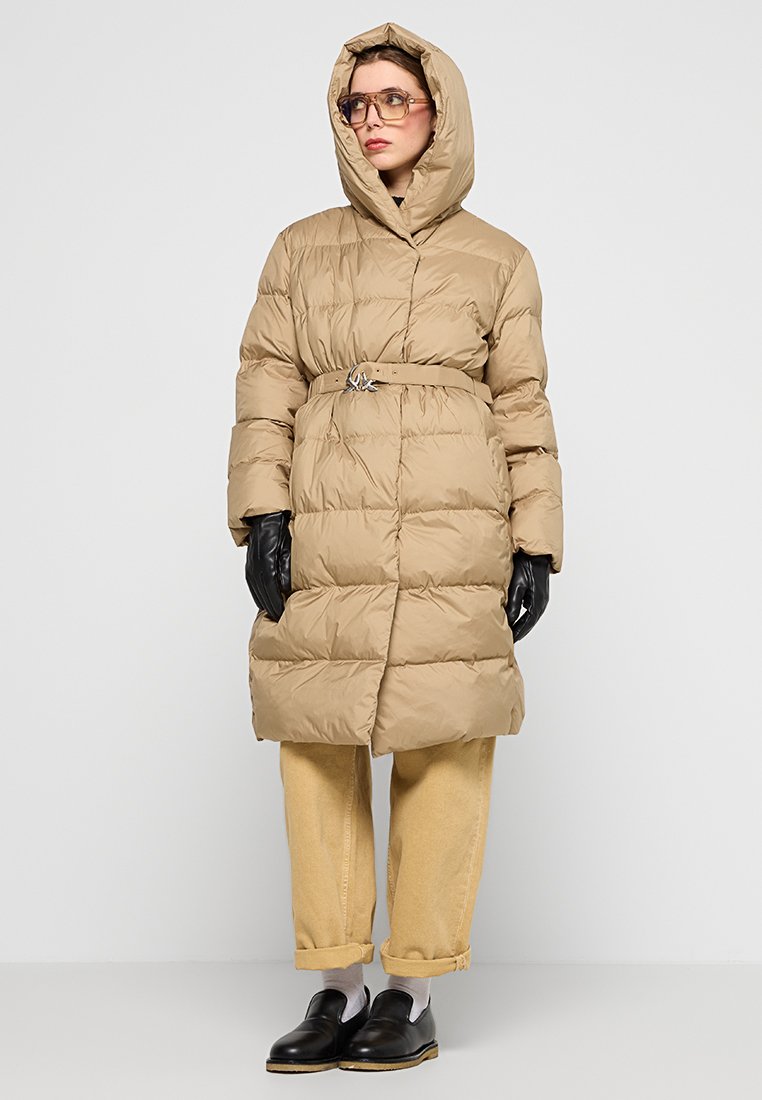Donna con un cappotto imbottito beige con cintura e cappuccio, guanti neri, pantaloni beige arrotolati, calzini bianchi, scarpe nere e occhiali grandi e oscurati.