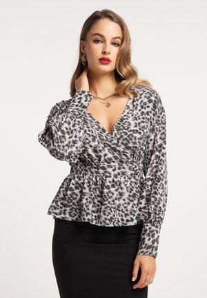 Frau mit welligem Haar, die eine graue und schwarze Leopardenmuster-Bluse mit Puffärmeln und einen schwarzen Rock trägt, posiert mit einer Hand nahe am Hals.