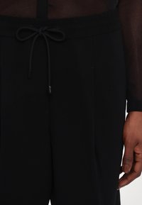 Černé kalhoty na stažení s tkaničkou, vyrobené z měkké látky, s páskem v pase a bočn�ími kapsami. Hladká textura s volným střihem.