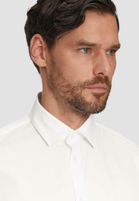 Witte katoenen blouse met een gestructureerde kraag, voorbies met knopen en een gladde textuur, gedragen door een man met kort haar en een baard.