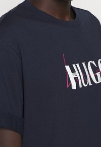 Granatowa bawełniana koszulka z krótkim rękawem z białym i różowym graficznym napisem "HUGO". Gładka faktura, okrągły dekolt.