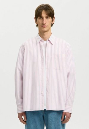 Młody mężczyzna ubrany w jasnoróżową i niebieską koszulę w paski zapinaną na guziki, na białej koszulce i niebieskich dżinsach, stojący na jednolitym tle.