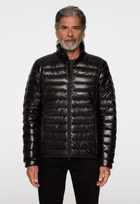 Svart pufferjacka med horisontellt quiltat mönster, hög krage och dragkedjestängning. Glansig finish och tät passform. Bärs över en svart skjorta.