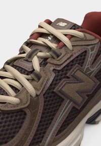 Καφέ και μπεζ αθλητικό παπούτσι New Balance με πλεκτό επάνω μέρος, επενδύσεις από σουέτ και μπεζ κορδόνια σε λευκή σόλα, κοντινή λήψη υπό γωνία.