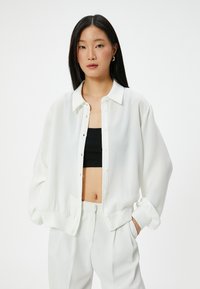 Camicia bianca con bottoni, dal taglio relax e dalla trama morbida. Presenta un colletto a punta e polsini elasticizzati. Indossata sopra un top nero.