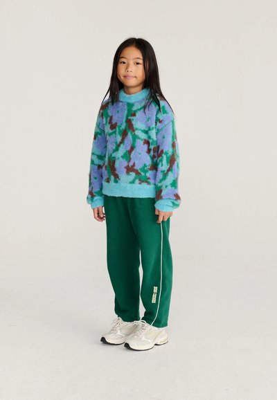 Jeune fille debout, portant un pull duveteux à motifs bleu-vert, un pantalon de jogging vert foncé avec une bande blanche, et des baskets blanches sur fond uni.