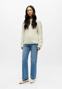 Cremfarbener Strickpullover mit langen Ärmeln und dekorativen Perlenakzenten, kombiniert mit hellblauen Schlagjeans und beigen Schuhen.