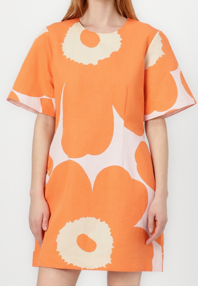 Femme portant une robe orange à manches courtes, au-dessus du genou, avec de grands motifs floraux blancs et beiges.