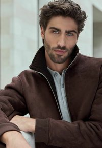 Jachetă din suede maro cu un guler texturat căptușit cu fleece. Are un fermoar în față și manșete răsucite. Sub aceasta se află o cămașă gri deschis.