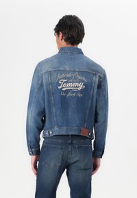 Jeansjakke i middels blå med en strukturert form, med en logo print på baksiden, knappelukking og et lærdetalj på kanten.