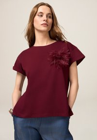 T-shirt bourgogne à manches courtes avec ourlets retroussés, présentant un détail floral texturé sur le côté gauche. Tissu en coton doux, coupe décontractée.