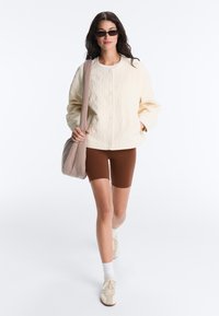 Femme marchant portant une veste matelassée crème, un short de vélo marron, des baskets blanches, des lunettes de soleil et portant un sac à bandoulière beige.