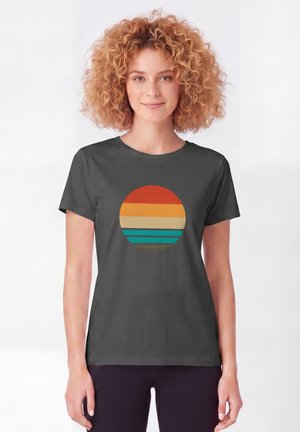 Jeune femme aux cheveux roux bouclés portant un t-shirt gris foncé avec un dessin circulaire de rayures horizontales aux couleurs du coucher de soleil.