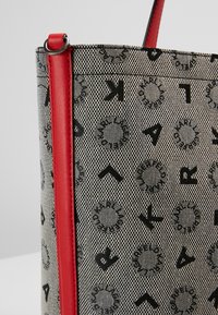 Sac à main gris texturé avec motifs circulaires et lettres noires, bande verticale en cuir rouge et anse rouge avec anneau métallique.