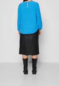 Blaues, strukturiertes Langarmshirt mit Schlüsselloch-Rücken-Detail, kombiniert mit einem schwarzen Leder-Bleistiftrock und kniehohen schwarzen Stiefeln.