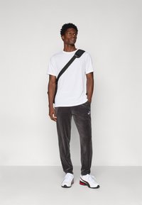 Biała koszulka z krótkim rękawem, szare dresowe spodnie z weluru z logo, czarna torba na ramię i białe sneakersy z czerwonymi akcentami. Prosty design i luźny krój.