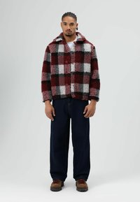 Rood-grijs geruite nep bontjas met kraag, knopen aan de voorkant, gecombineerd met donkere denim broek en bruine laarzen. Gladde textuur.