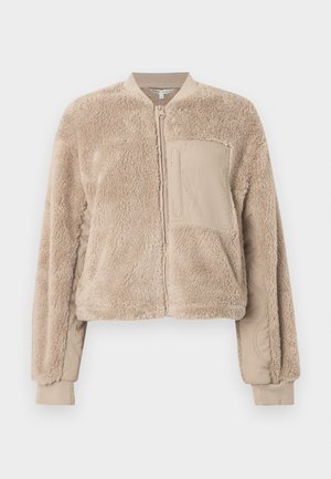 Veste aviateur beige avec une texture douce et duveteuse sur le corps et un tissu lisse sur les manches. Comprend une poche avant et des poignets côtelés.