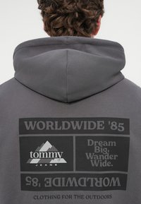 Γκρίζα βαμβακερή φούτερ με τύπωμα μπροστά που περιλαμβάνει "WORLDWIDE '85", "tommy JEANS" και ένα λογότυπο βουνού. Με κουκούλα και υφασμάτινη υφή.