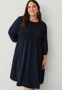 Købmandsblå kjole med en smocked bodice, lange pufærmer og en løs, flydende nederdel. Lavet af blødt stof med en glat overflade.