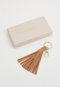 Tiroir en cuir tan avec un pompon, muni d'un mousqueton en métal doré, accompagné d'une boîte rose clair portant le nom "Coccinelle" en lettres dorées.