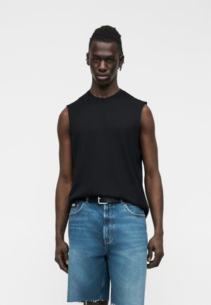 Fiatal, sötét bőrű férfi ujjatlan fekete pólóban és kék farmer rövidnadrágban, sima világos háttér előtt áll.