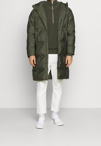 Olivgrüne Steppjacke mit Kapuze, mit Reißverschluss auf der Vorderseite, Brusttasche und strukturiertem Stoff. Kombiniert mit einem dunkelgrünen Pullover und weißen Hosen.