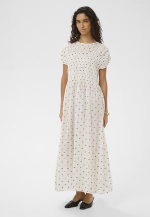 Woman with curly hair wearing a white ankle-length dress with small black patterns, puff sleeves, and black flat shoes, standing on white background.