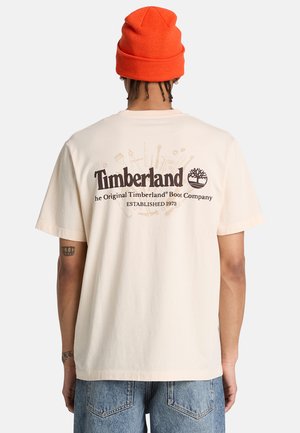 Persona che indossa una t-shirt Timberland beige con il testo del logo sul retro, abbinata a jeans blu e un berretto arancione.