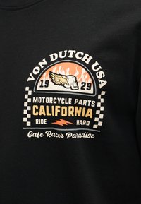 Σχέδιο μαύρου πουκαμίσου με κρανίο με φτερά, φλόγες, καρό μοτίβο και κείμενο που αναφέρεται σε "Von Dutch USA", "Motorcycle Parts California" και "Ride Hard".