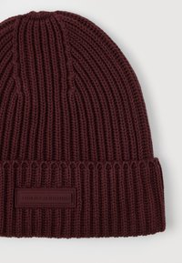 Ciemnobrązowa, prążkowana czapka beanie z podwiniętym mankietem i prostokątną łatką logo Tommy Hilfiger z przodu, na jasnym tle.
