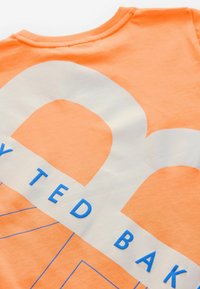 Oranžs kokvilnas T-krekls ar apaļu kaklu, ar lielu baltu un zilu grafisko dizainu un zilu uzrakstu "TED BAKER."