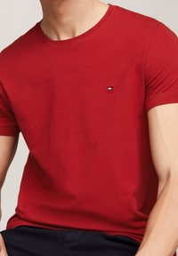 Rød bomulds T-shirt med rund hals og korte ærmer, der har et lille broderet logo på venstre bryst. Glat tekstur.