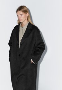 Cappotto oversize in lana nera con rever a punta, due bottoni e tasche laterali. Indossato sopra una camicia a bottoni grigia chiara. Texture liscia.