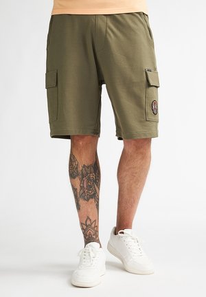 Olivgrüne Cargo-Shorts mit zwei Seitentaschen, einem dekorativen Aufnäher und einer weichen Textur. Kombiniert mit weißen Sneakers.