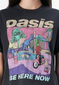 T-shirt nero con un'illustrazione grafica colorata che presenta il testo "oasis" sopra e "BE HERE NOW" sotto, raffigurante una scena con persone e veicoli d'epoca.