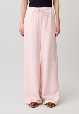 Mujer vestida con pantalones anchos de color rosa claro con cintura ajustable con cordón y zapatos marrones, de pie frente a un fondo claro liso.