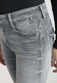 Jean en denim gris avec une coupe slim, comportant une poche sur la jambe droite, une fermeture à bouton métallique et une texture légèrement usée.