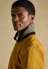 Jonge man met kort krullend haar, gekleed in een mosterdgele jas met een opstaande geruite kraag, kijkt zelfverzekerd opzij tegen een effen achtergrond.