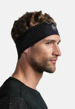 Hombre con cabello corto, rizado y castaño lleva una cinta deportiva negra y una camiseta deportiva negra, mirando hacia la derecha con expresión neutral.