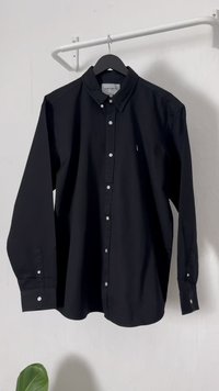 Camisa de manga longa preta, com botões e feita de algodão, apresentando um colarinho clássico, botões brancos e um pequeno logotipo no peito.