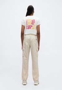 Γυναίκα όρθια με την πλάτη στραμμένη, φοράει λευκό t-shirt με ροζ και κίτρινο λουλουδένιο σχέδιο και μπεζ παντελόνι cargo με αθλητικά παπούτσια.